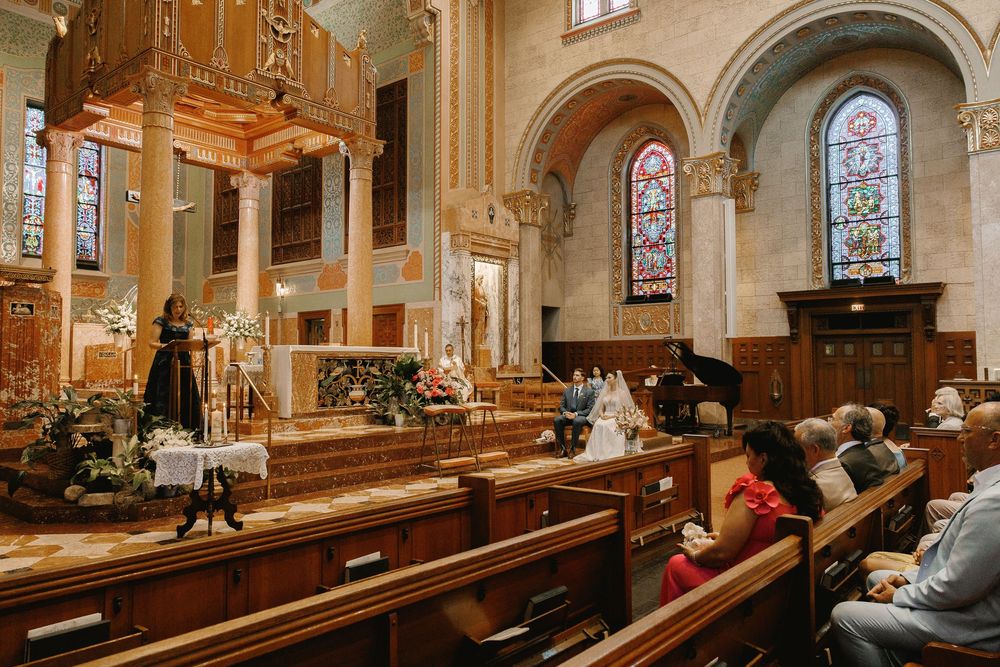 Guests seated in wooden church pews during an elegant wedding ceremony with stained glass windows.