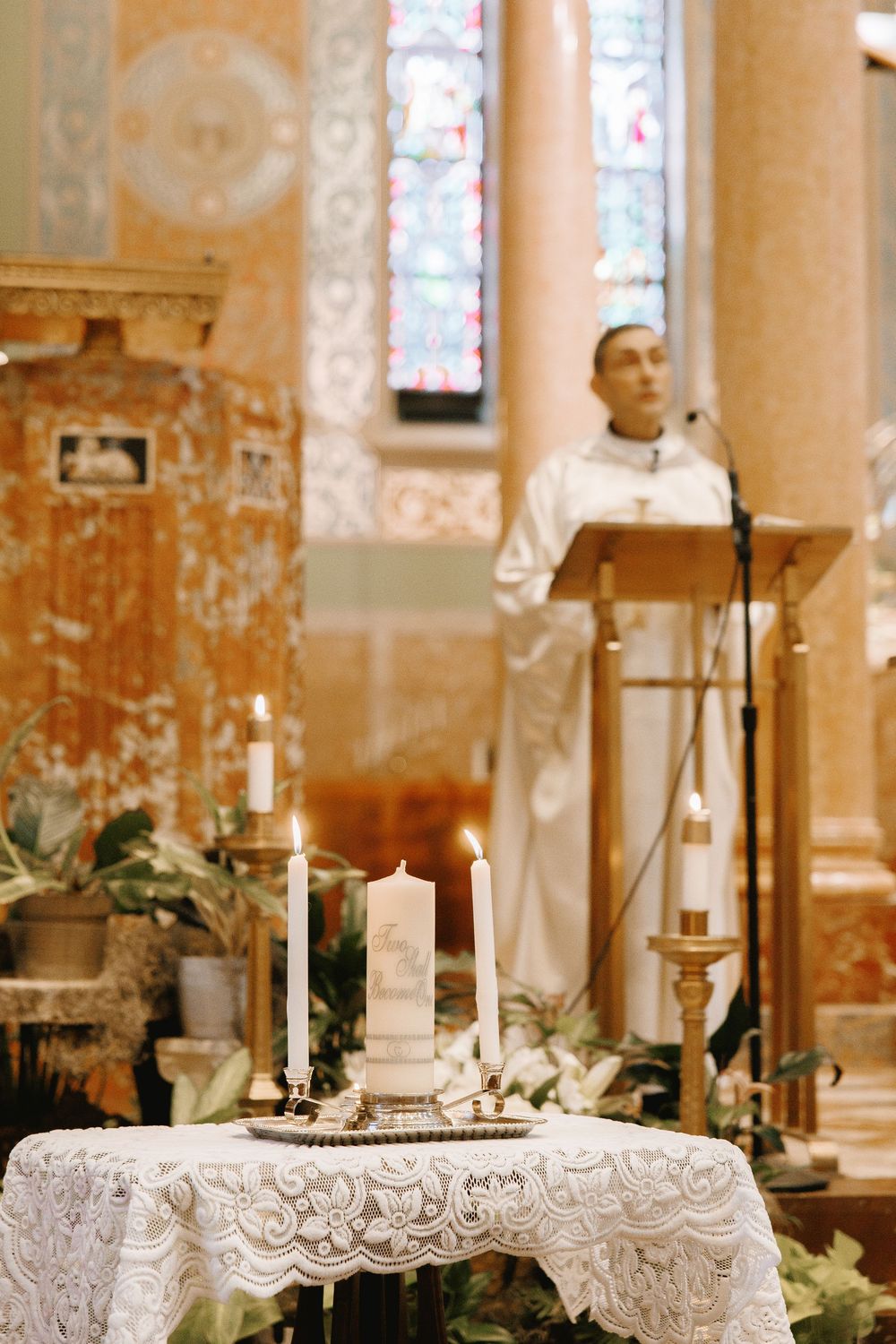 Priest at podium during church wedding ceremony with lit candles on lace-covered altar table in ornate chapel.