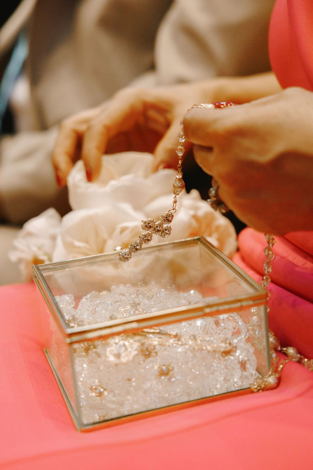 Hands holding a delicate crystal rosary over a glass box filled with pearls at a wedding ceremony.