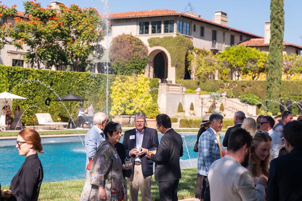 Guests mingle poolside at an elegant mansion garden party with a fountain and lush landscaping on a sunny day.