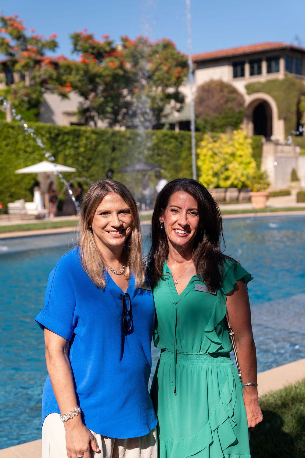Two women smiling outdoors near a fountain, one in blue blouse and one in green dress, with a building and trees behind.