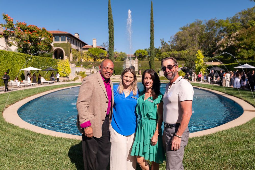 Four smiling people pose together in front of an elegant fountain and lush garden at a sunny outdoor event.