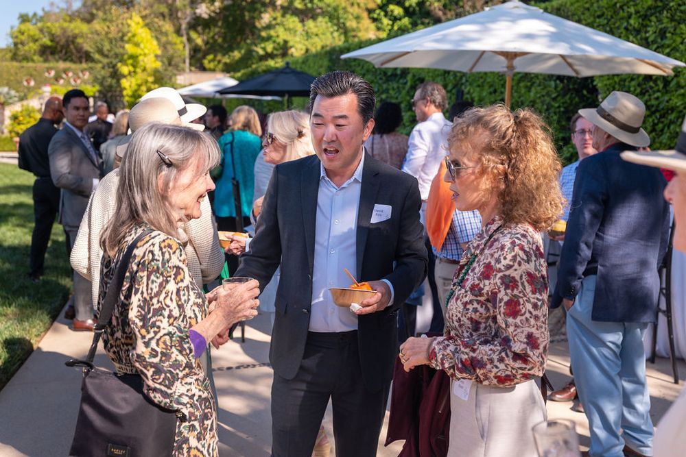 Three professionals networking at an outdoor social gathering event under a patio umbrella on a sunny day.