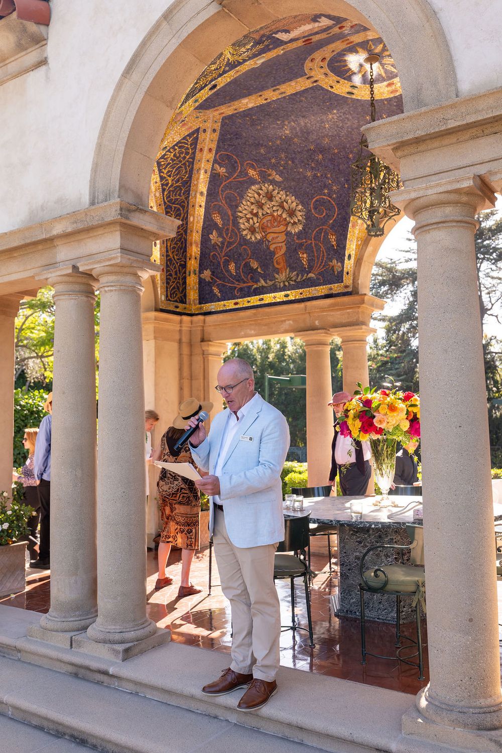 Man in white blazer reads at ornate archway with mosaic ceiling, columns, and floral arrangement at elegant outdoor venue.