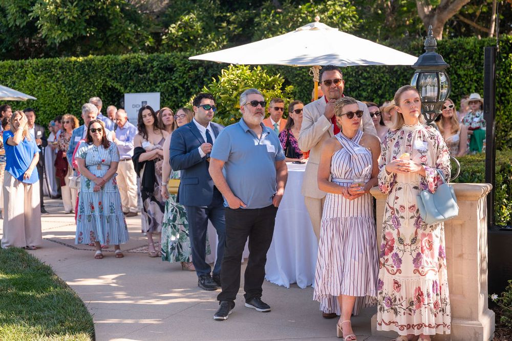 Elegantly dressed guests line up outdoors at a sunny garden event, socializing along a manicured pathway.