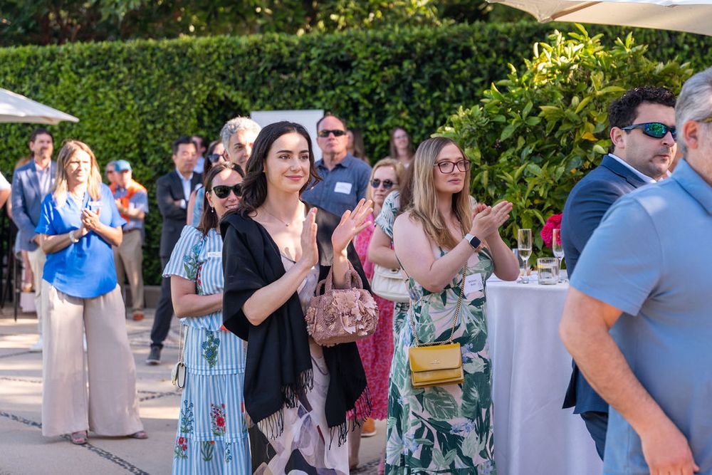 Guests applauding at an outdoor garden party event, dressed in summer attire, with lush greenery in the background.
