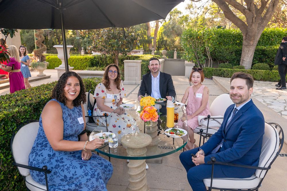 Five professionals smile around a glass patio table at an outdoor garden event, dressed in business casual attire.