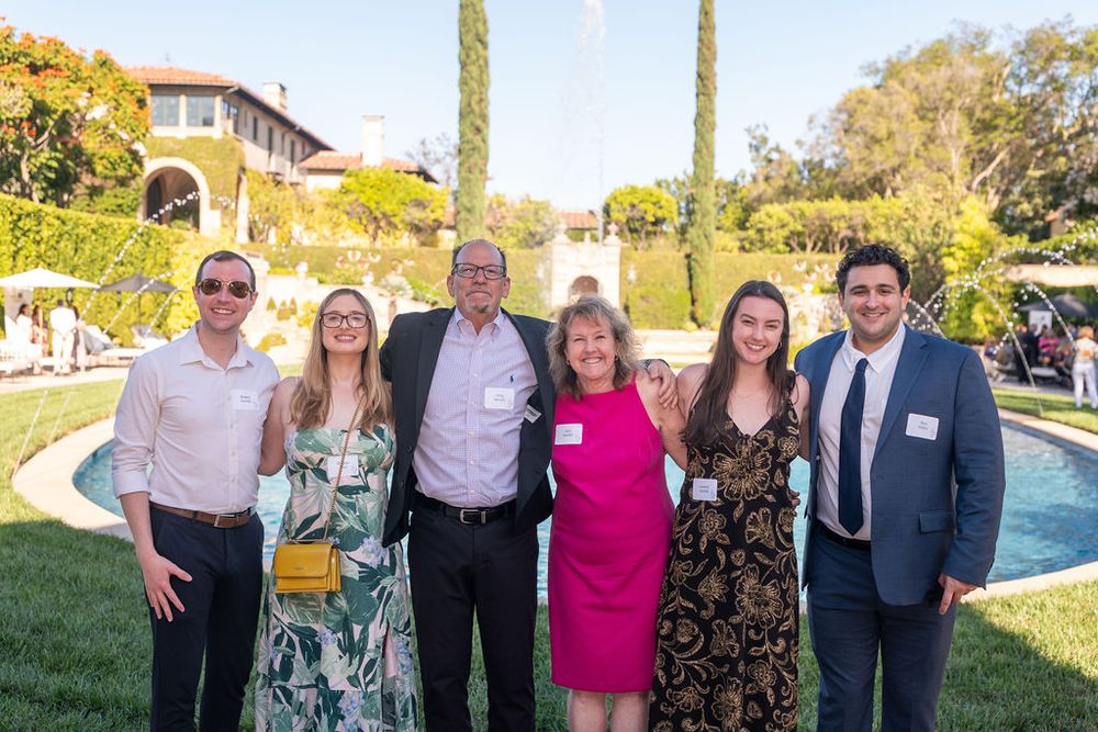 Six people smiling together outdoors at an elegant garden party with a fountain and lush greenery in the background.