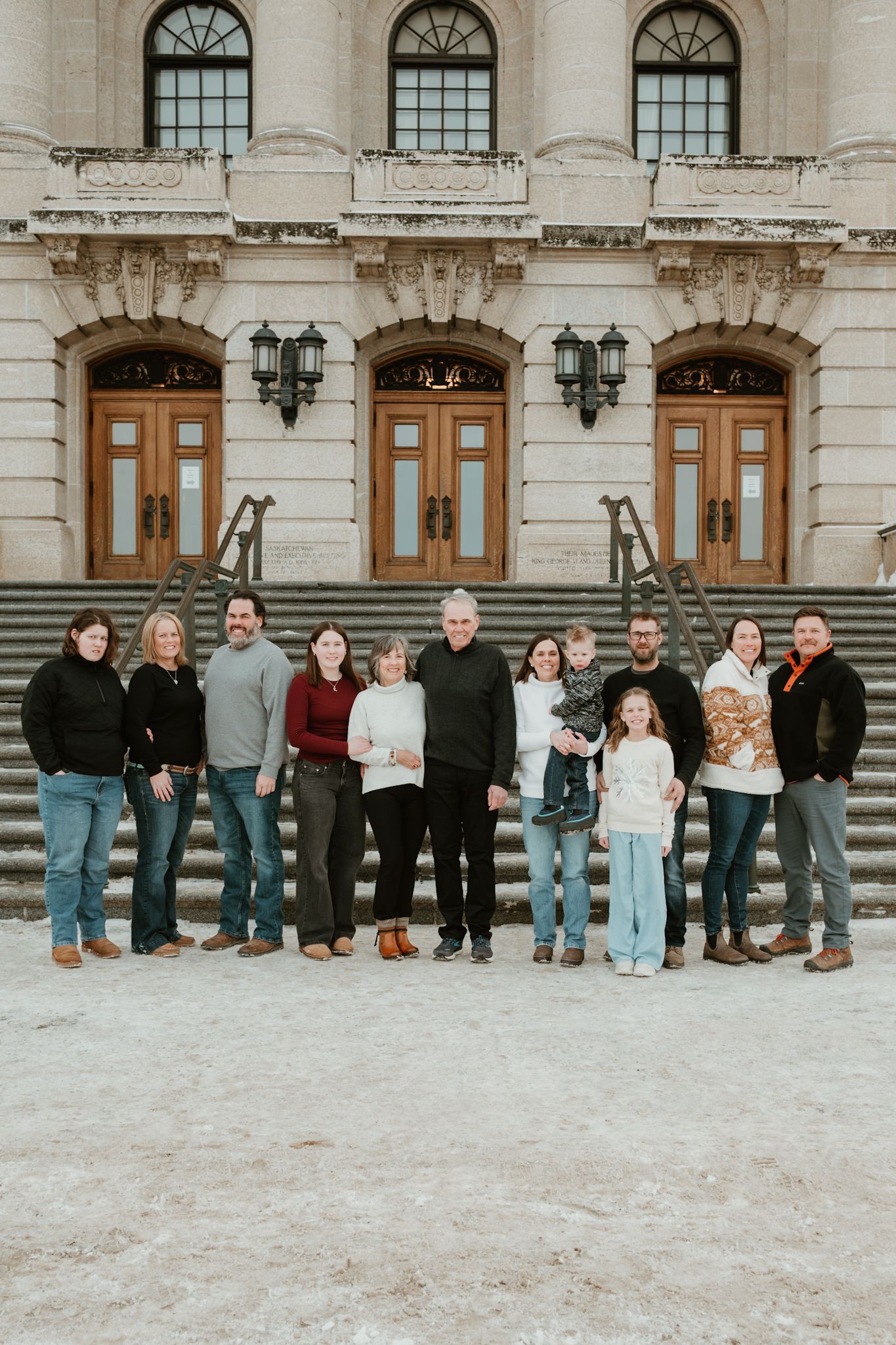 Large family group portrait posed on snowy steps of a grand historic building with arched windows and wooden doors.