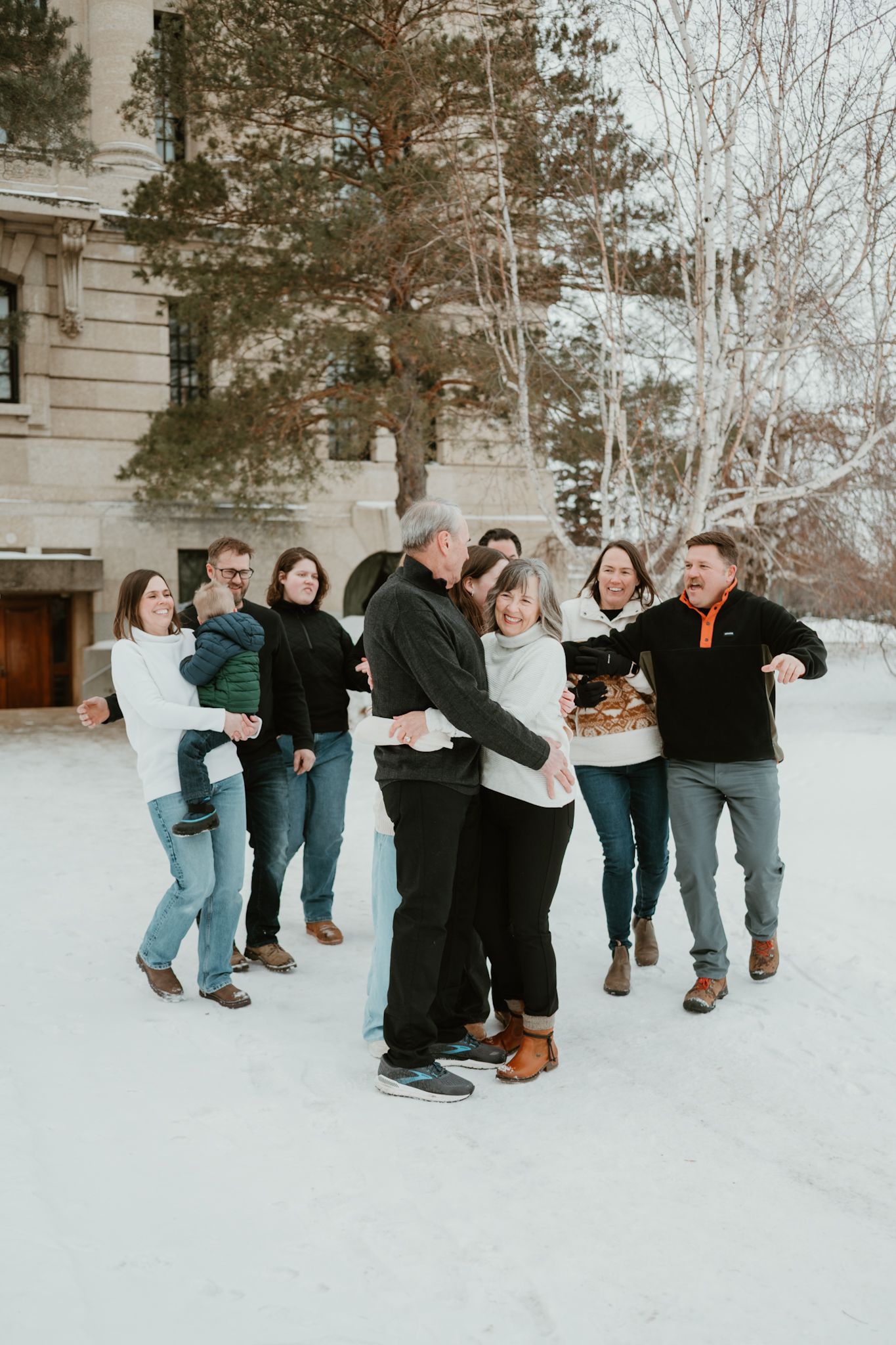 Family group poses outdoors in snow near historic building, central couple embraces while others celebrate around them in winter attire.