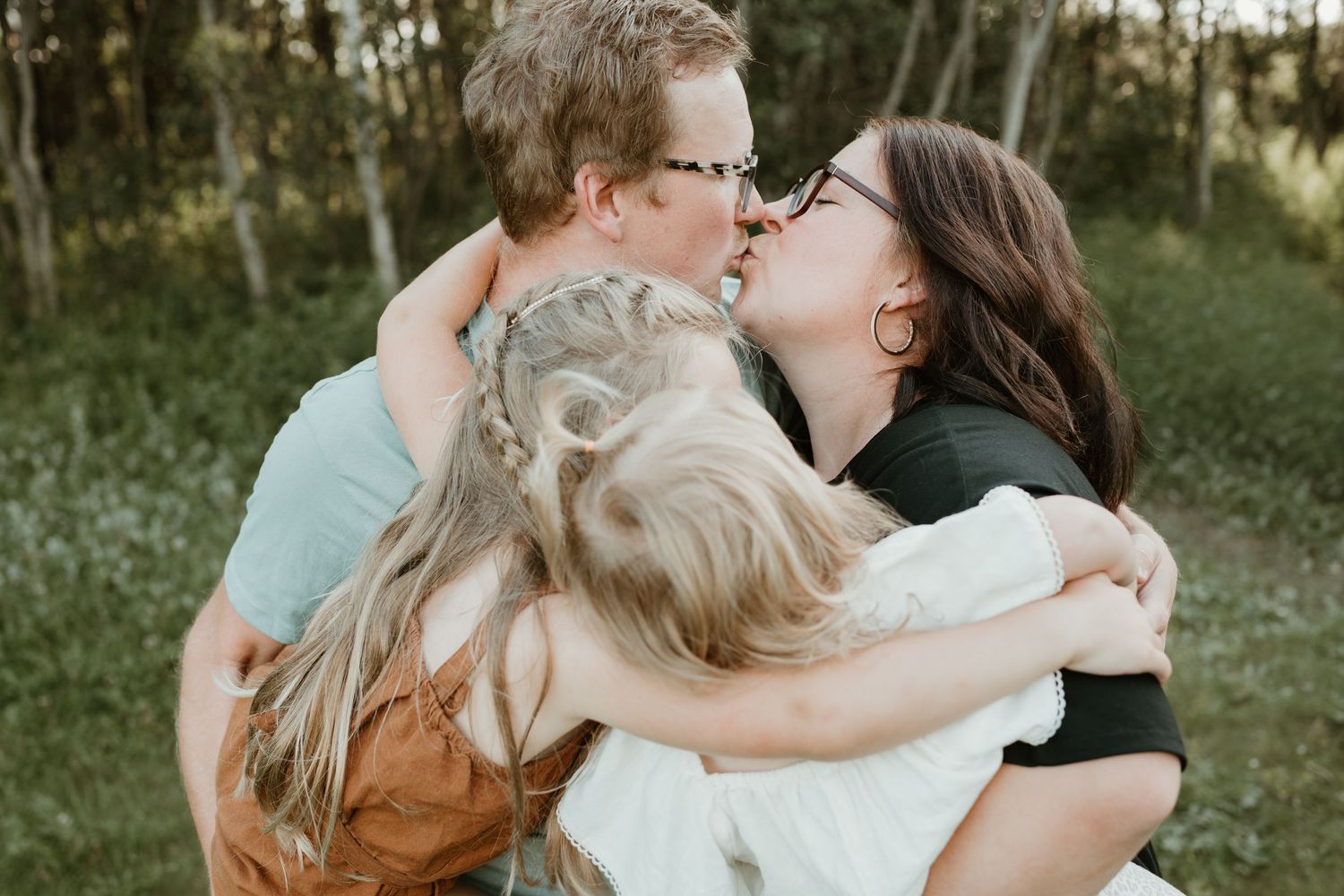 A couple shares a kiss while embracing two young children in a warm family hug outdoors.