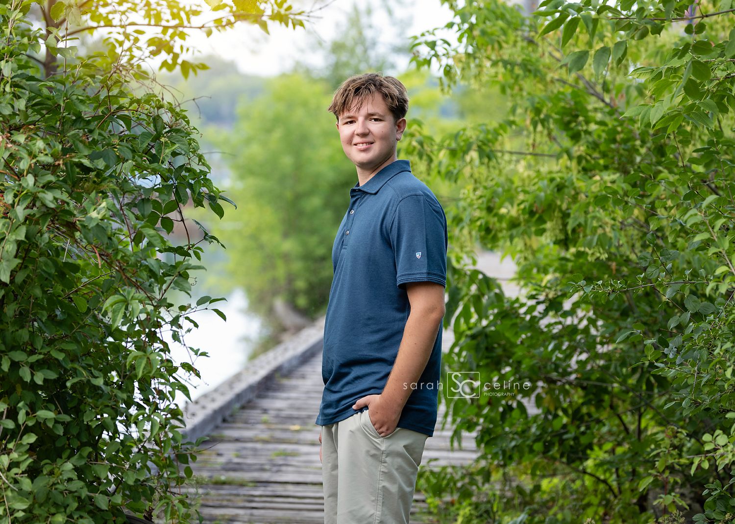 Smiling teenage boy in blue polo shirt standing on wooden boardwalk surrounded by lush green trees near water.