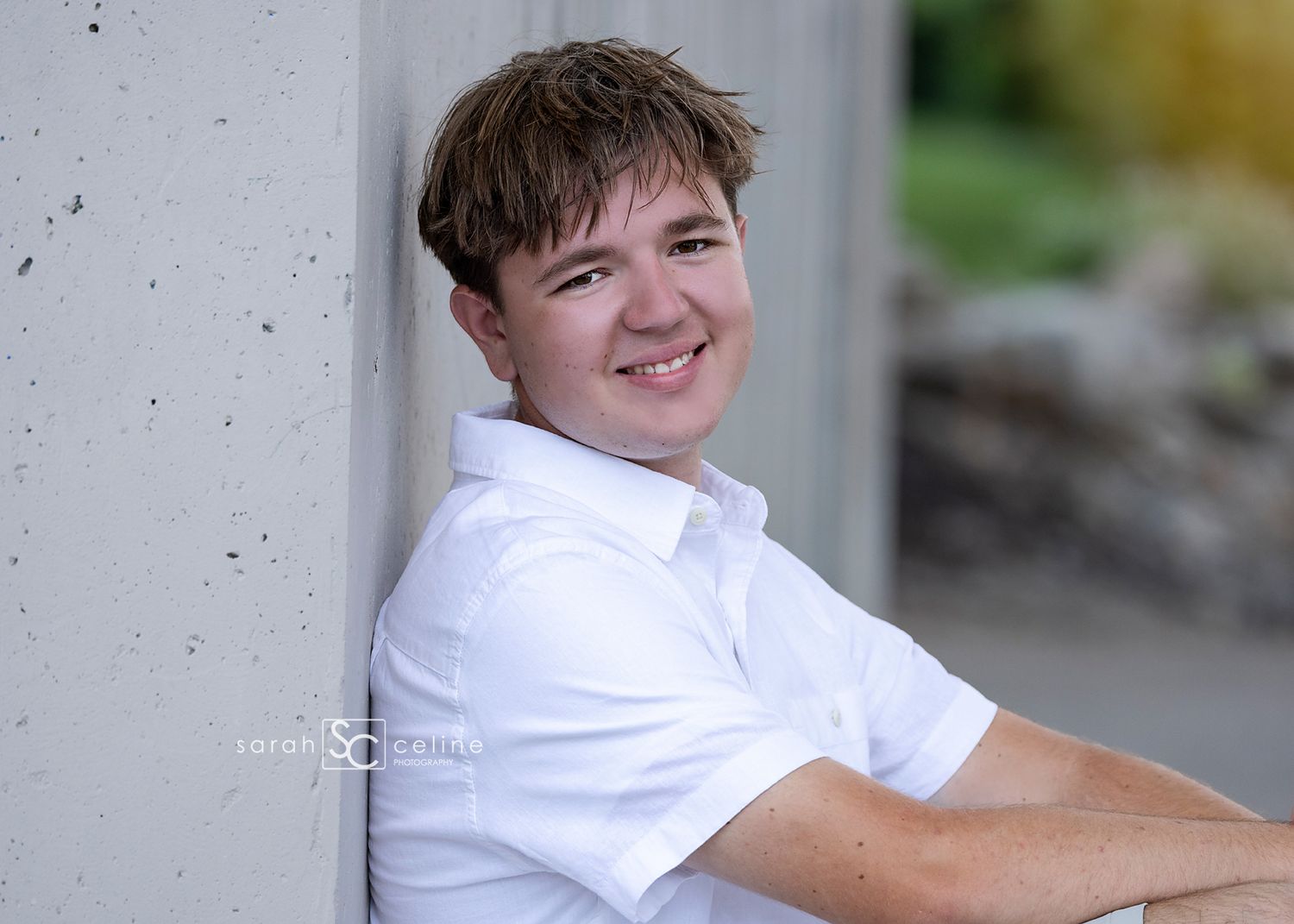 Smiling young man in white polo shirt leaning against concrete wall outdoors in natural light.