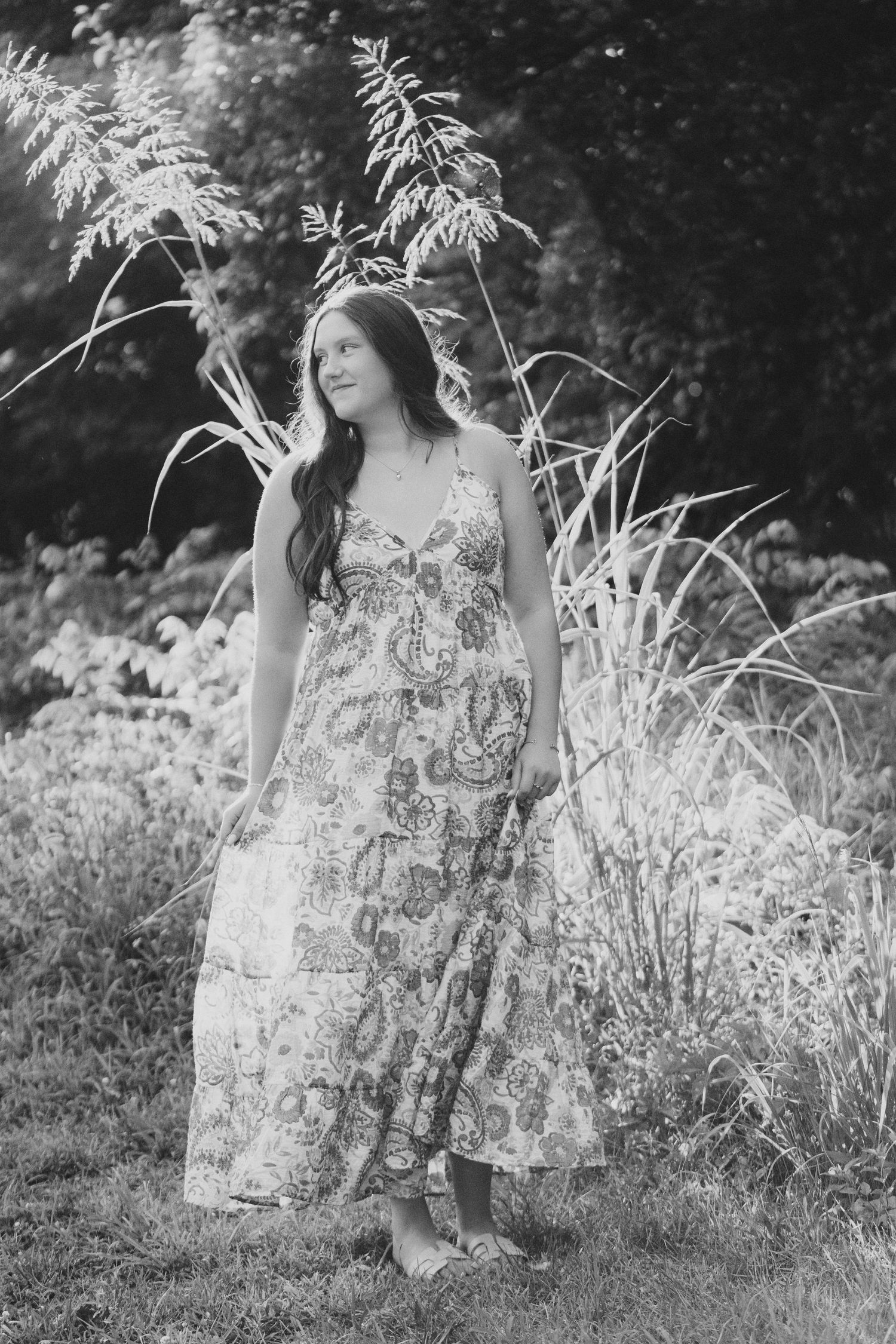 Black and white photo of a young woman in a floral maxi dress standing in a lush garden.
