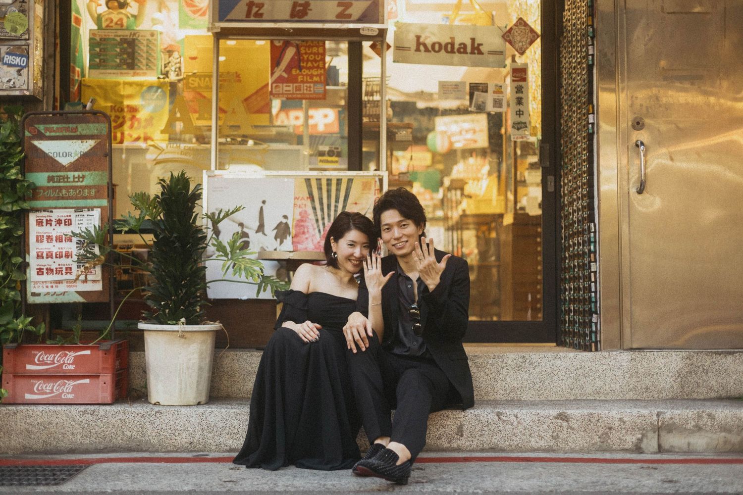 A smiling couple in white outfits sitting on vintage shop steps surrounded by plants and old utility meters.