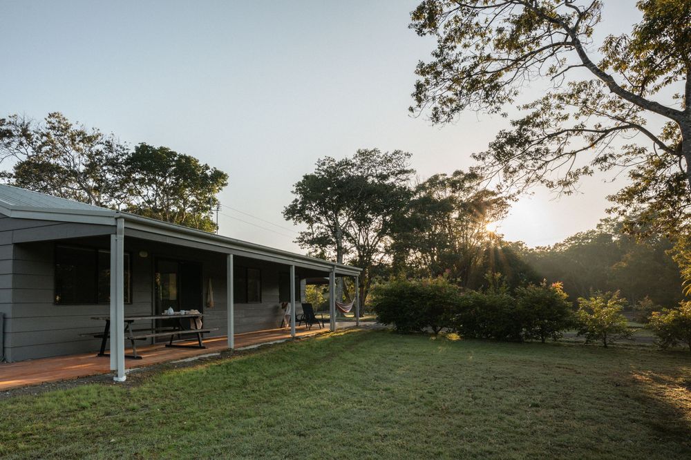 Rural homestead with covered veranda surrounded by lush green lawn and tall trees at golden sunset.
