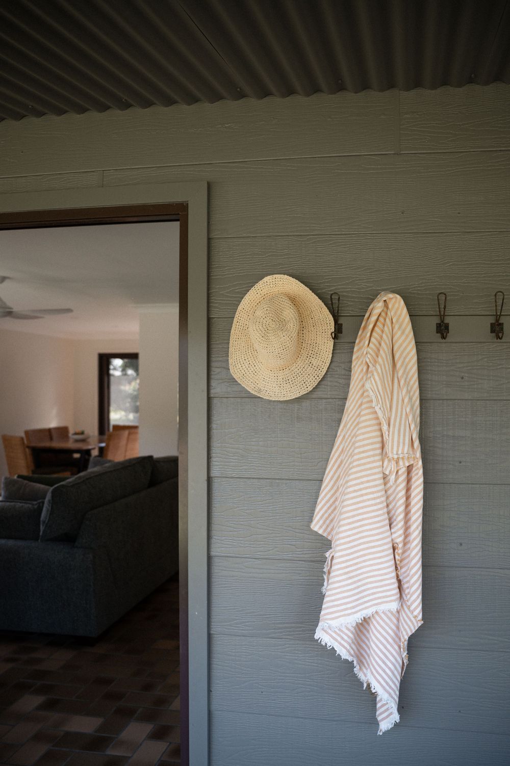 Straw hat and striped towel hanging on hooks beside a doorway of a grey timber coastal cottage interior.