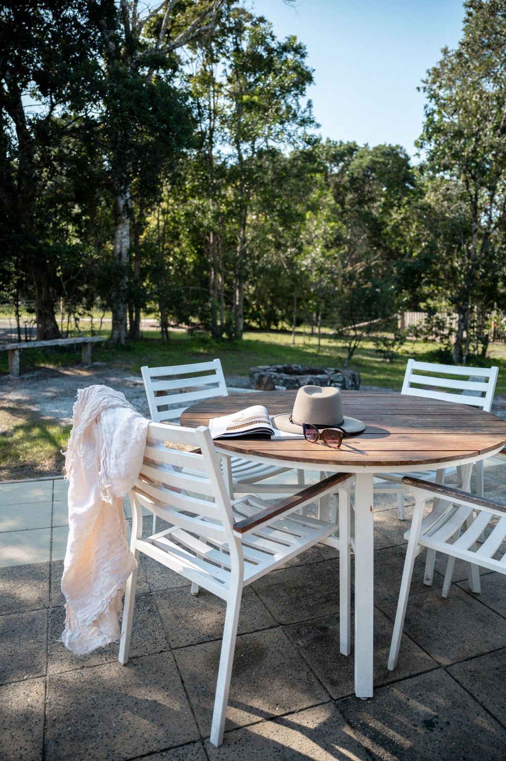 White outdoor dining set with teak table, open book, hat, and draped scarf on a patio surrounded by lush green trees.