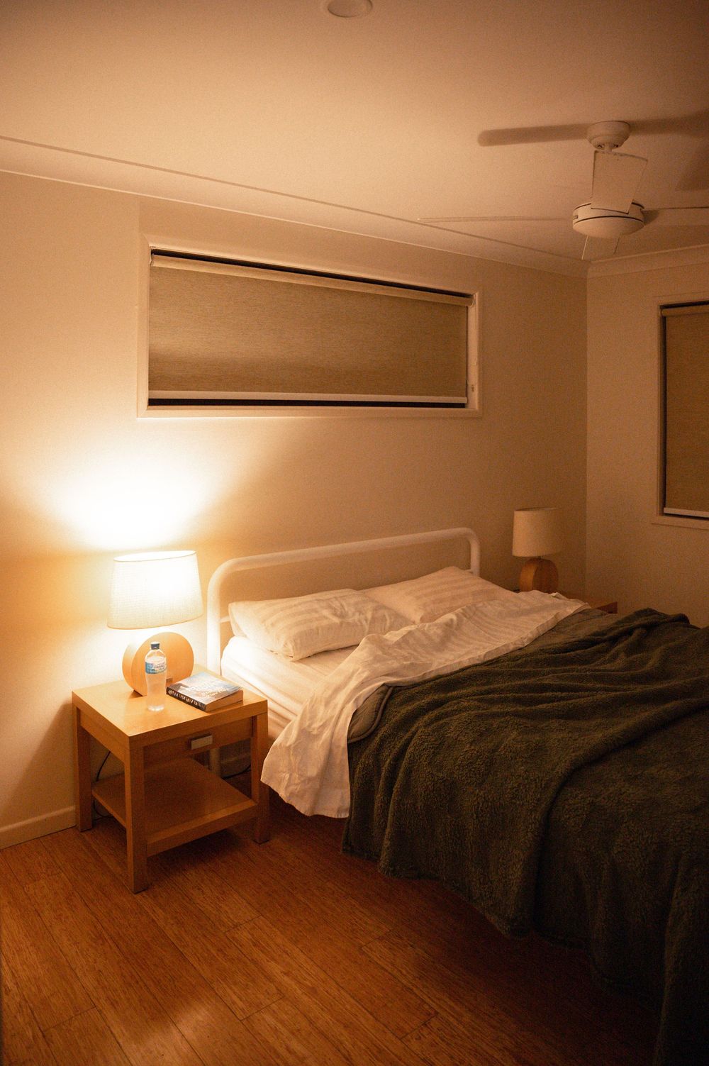 Cozy warmly lit bedroom with green blanket, wooden nightstand, lamp, and ceiling fan on hardwood floor.