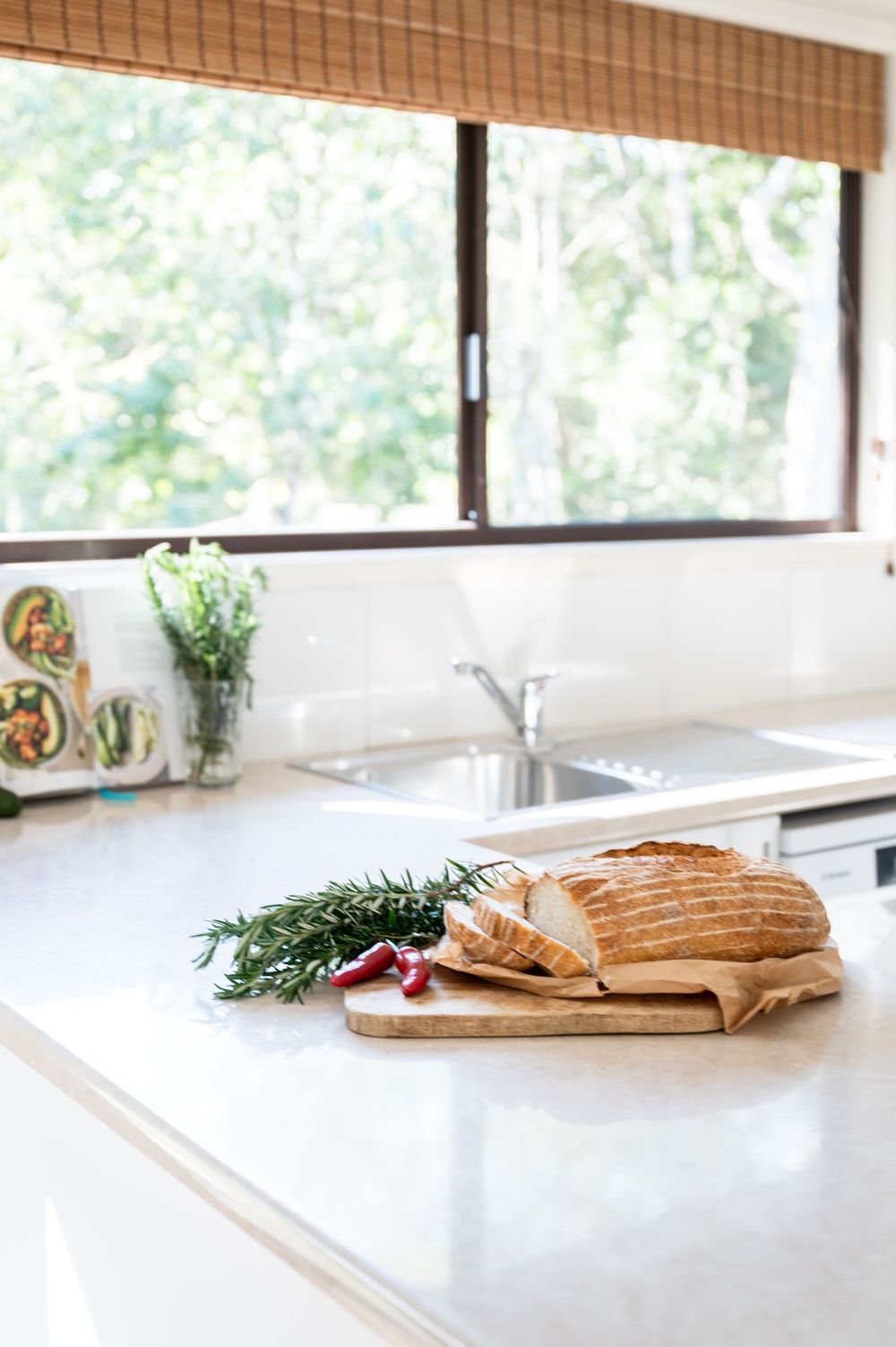 Sliced bread on wooden board with rosemary and chili on white kitchen counter near sink and window.