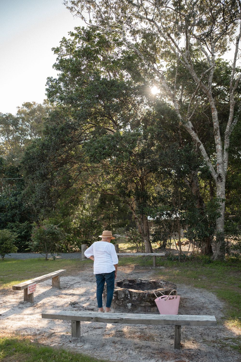 Woman in straw hat standing by rustic fire pit surrounded by wooden benches in lush garden setting.