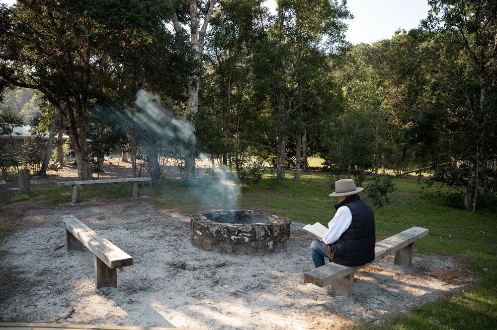 Person in cowboy hat sitting on wooden bench by a campfire in a wooded outdoor setting.