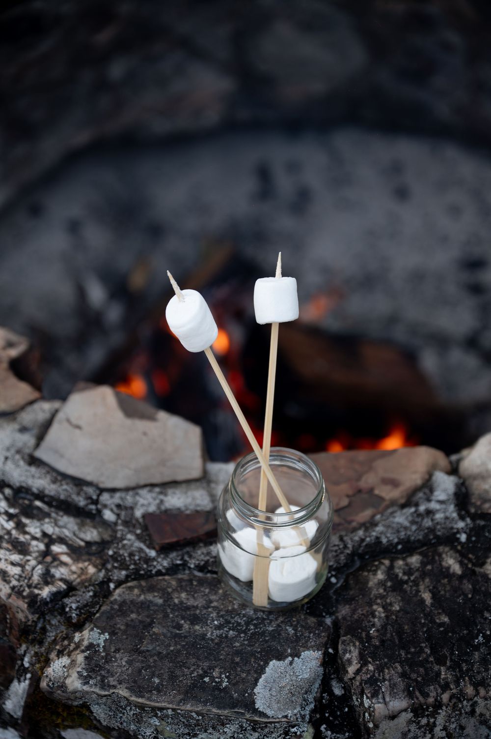 Two marshmallows on skewers resting in a glass jar beside a glowing campfire on rocky stones.