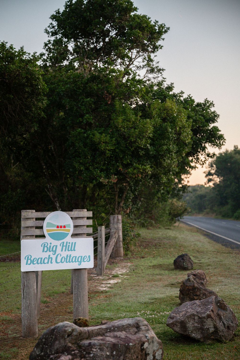 Entrance sign for Big Hill Beach Cottages beside a quiet road at dusk with trees.