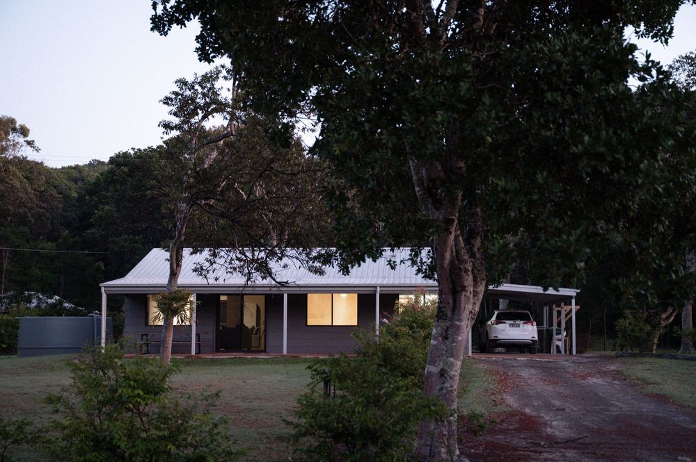 Charming rural house at dusk surrounded by tall trees with warm interior lights and a carport with vehicle.