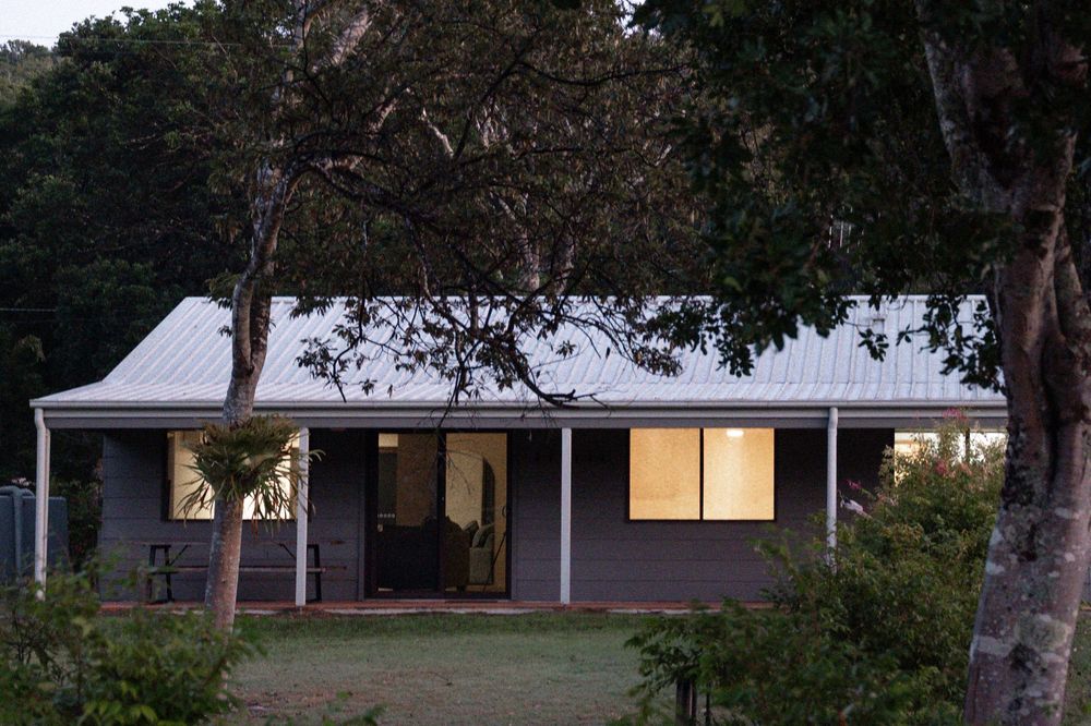 A charming single-story home with a white metal roof, covered veranda, and warm interior lighting surrounded by lush trees at dusk.