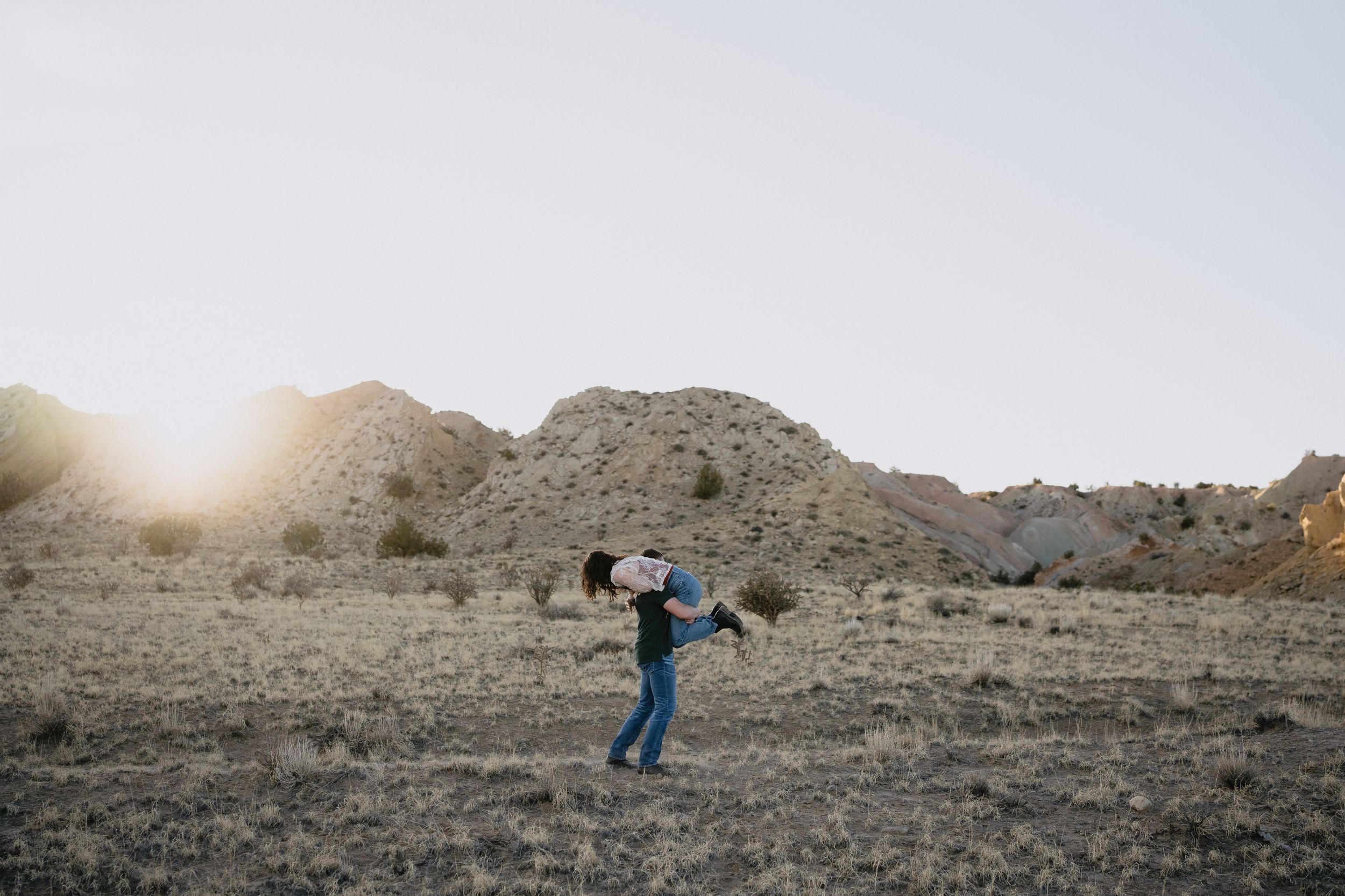 A person joyfully lifts a child in a vast desert landscape with rocky hills and golden sunlight in the background.