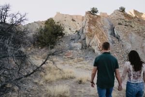 Couple holding hands walking toward dramatic sandstone cliffs in a desert landscape with sparse vegetation.
