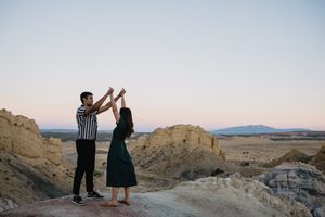 Couple dancing together on rocky desert landscape at sunset with sandy cliffs and mountains in background.