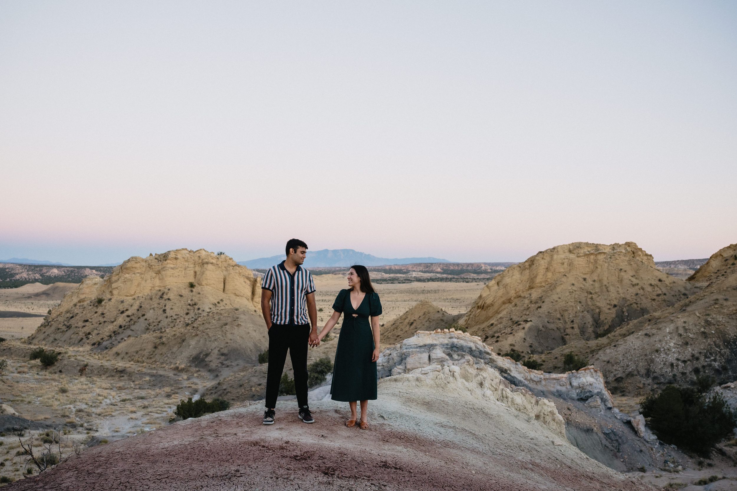 Couple holds hands on rocky desert landscape at dusk with sandy badlands formations and pastel sky behind them.