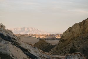 Arid desert landscape with layered rock formations framing a distant mountain range under a soft pastel sky.