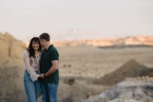 Couple smiling and holding hands in a desert landscape at golden hour with mountains in the background.