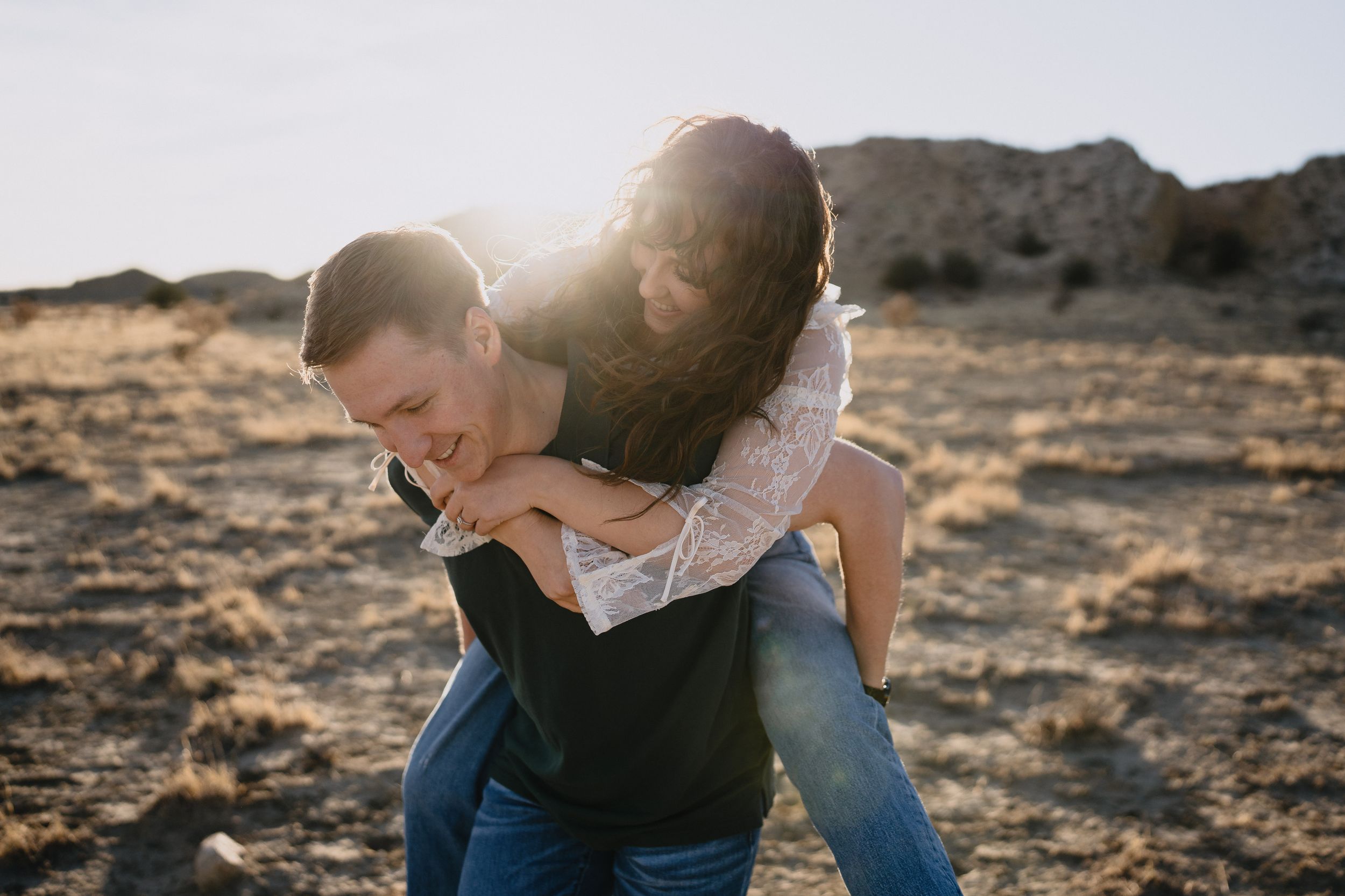 Couple laughing outdoors, woman piggyback riding man in sunny desert landscape.
