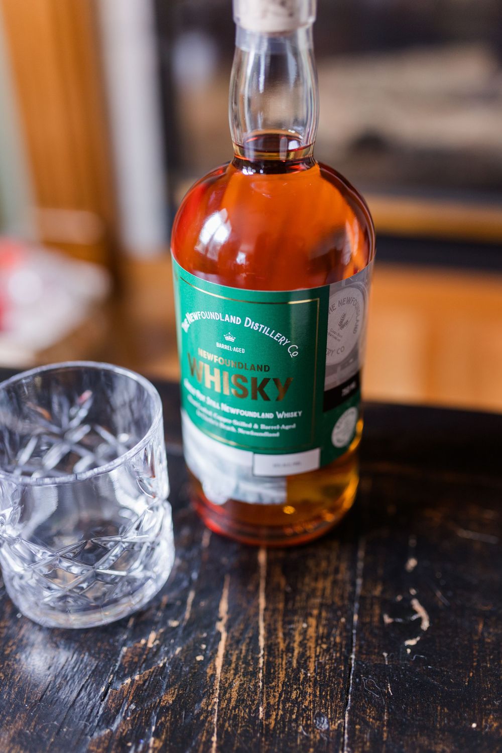 Newfoundland Distillery Co. whisky bottle with green label beside a crystal glass on a dark wooden table.