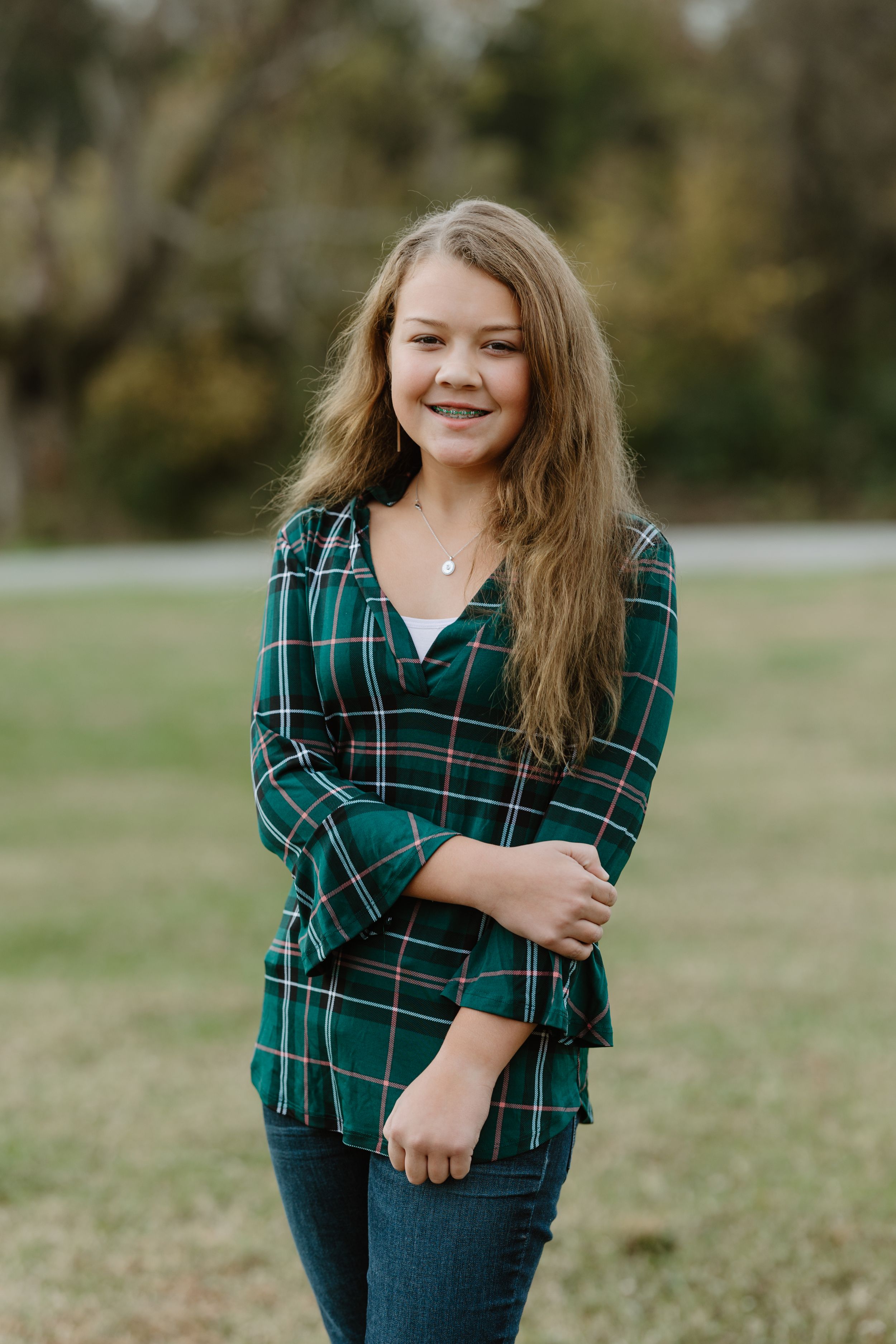 Young woman smiling in green plaid flannel shirt and jeans, posing outdoors in a grassy park setting.