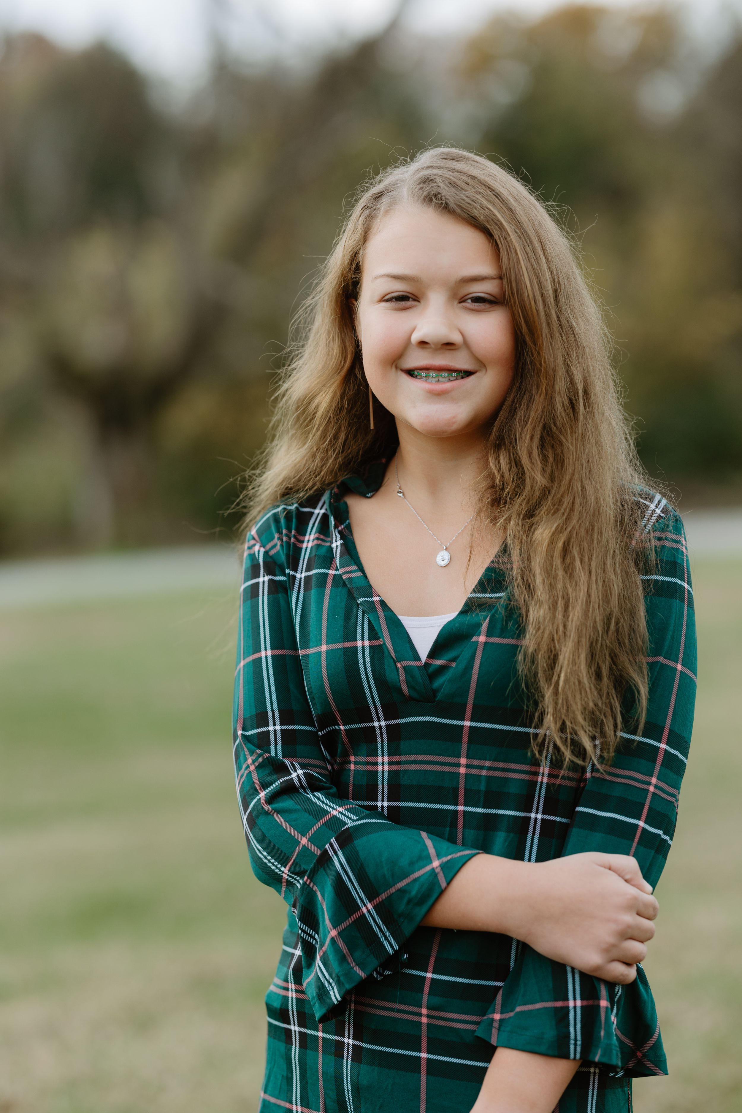 Smiling teenage girl with braces wearing green plaid shirt, long blonde hair, posing outdoors in park setting.