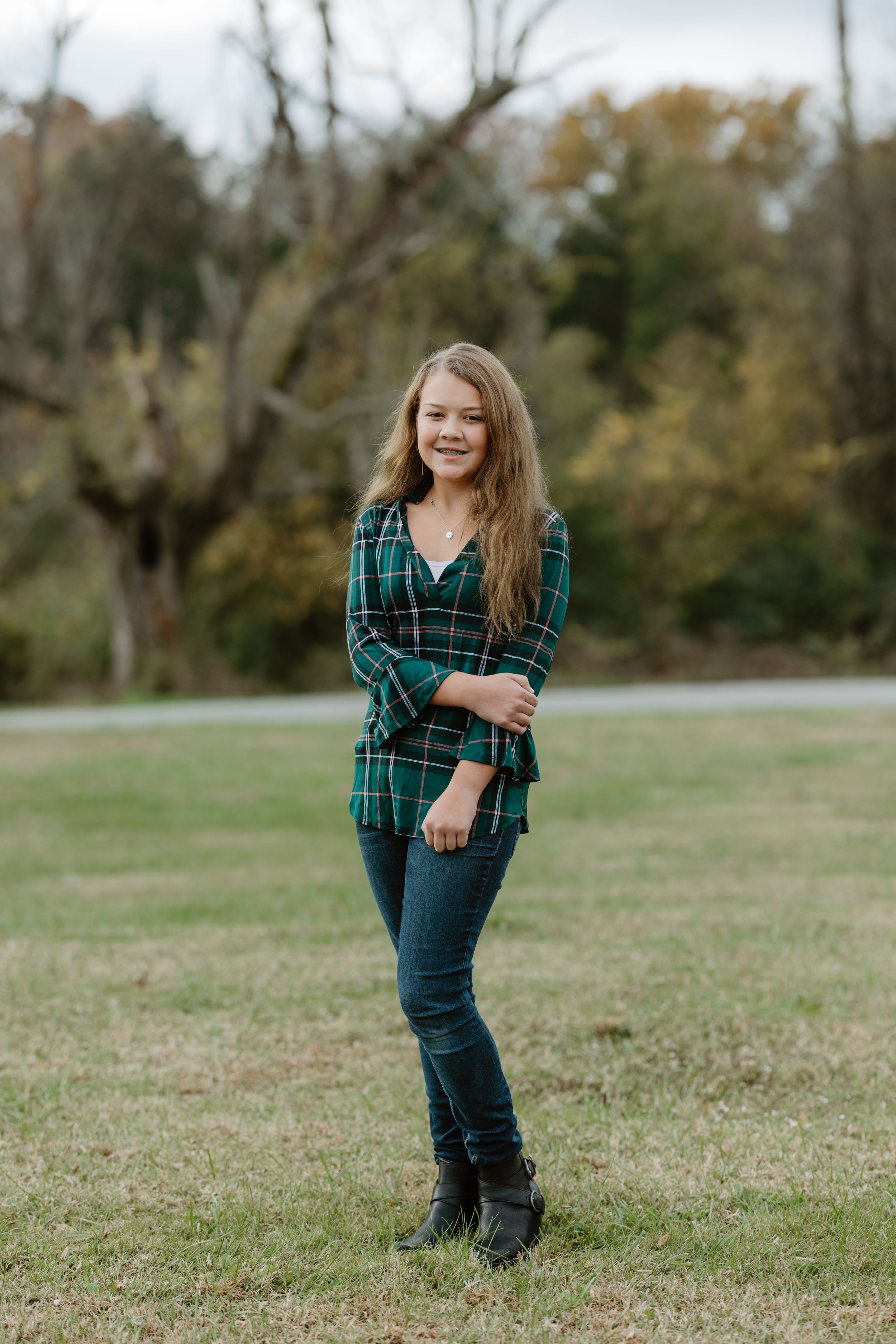 Young woman in green plaid shirt and jeans smiling outdoors in grassy field with autumn trees in background.