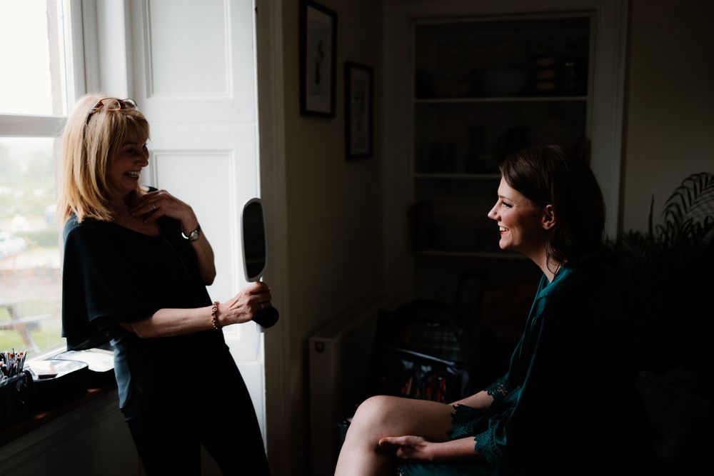 Two women engaged in conversation near a bright window in a dimly lit room, one standing in black dress.
