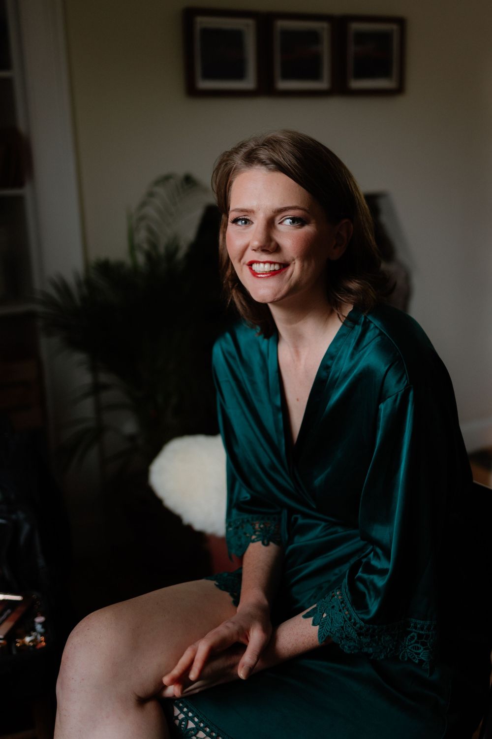 A smiling woman in a teal satin lace robe sits elegantly in a softly lit room, perfect for bridal getting-ready photos.