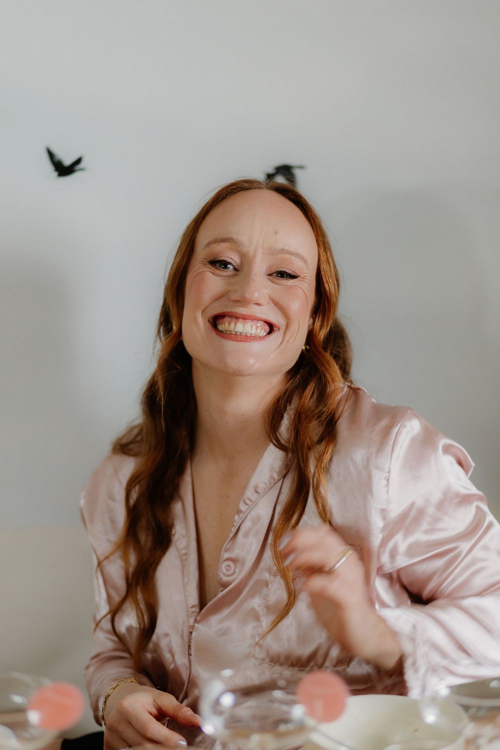 Smiling redhead woman in pink satin pajamas laughing joyfully at a table with confetti.