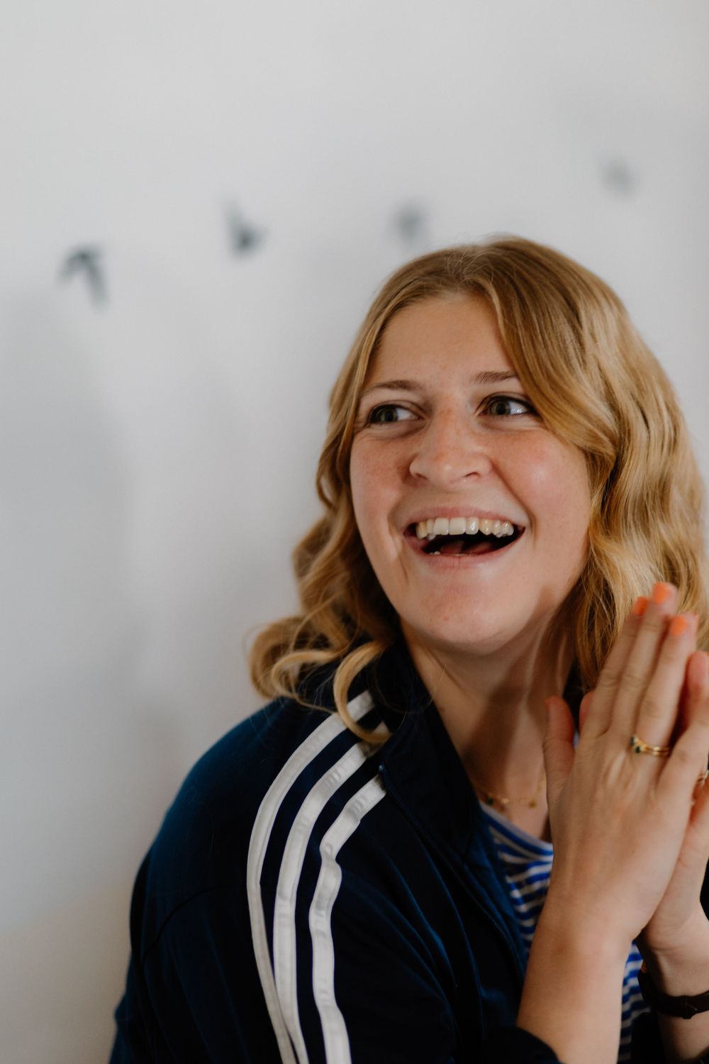 Laughing blonde woman in navy Adidas jacket clapping hands against white wall with bird decorations.