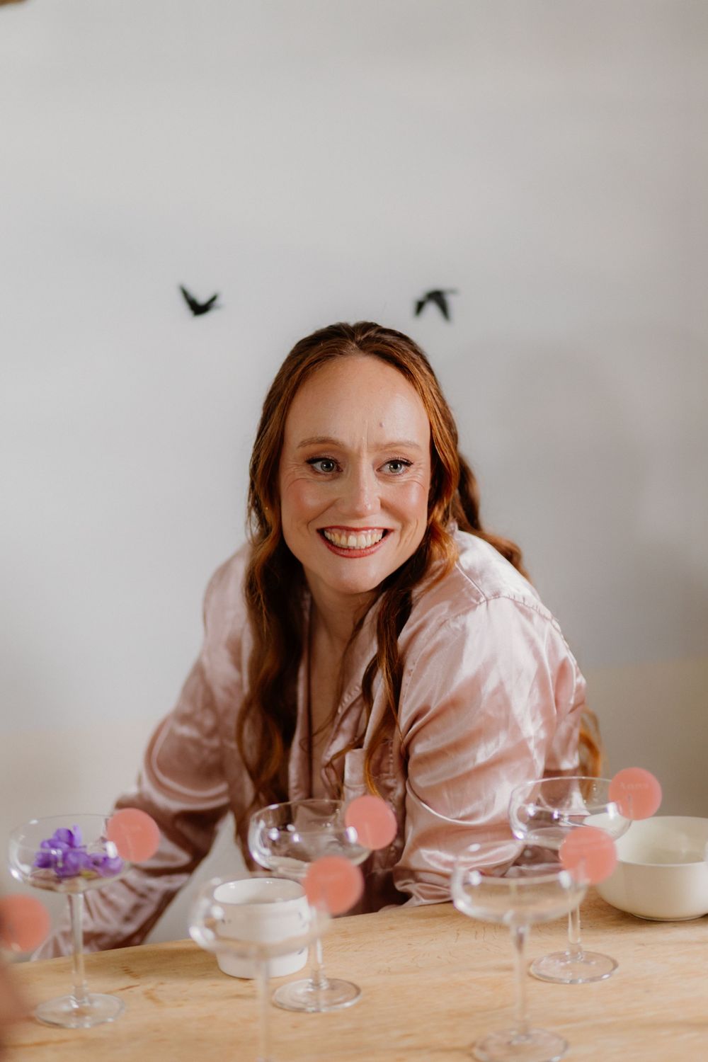 Smiling redhead woman in pink satin robe seated at table with elegant coupe glasses and floral decorations.