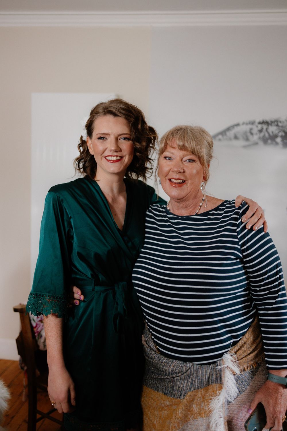 Two women smiling together, one in a green satin robe and one in a navy striped top, posing indoors.