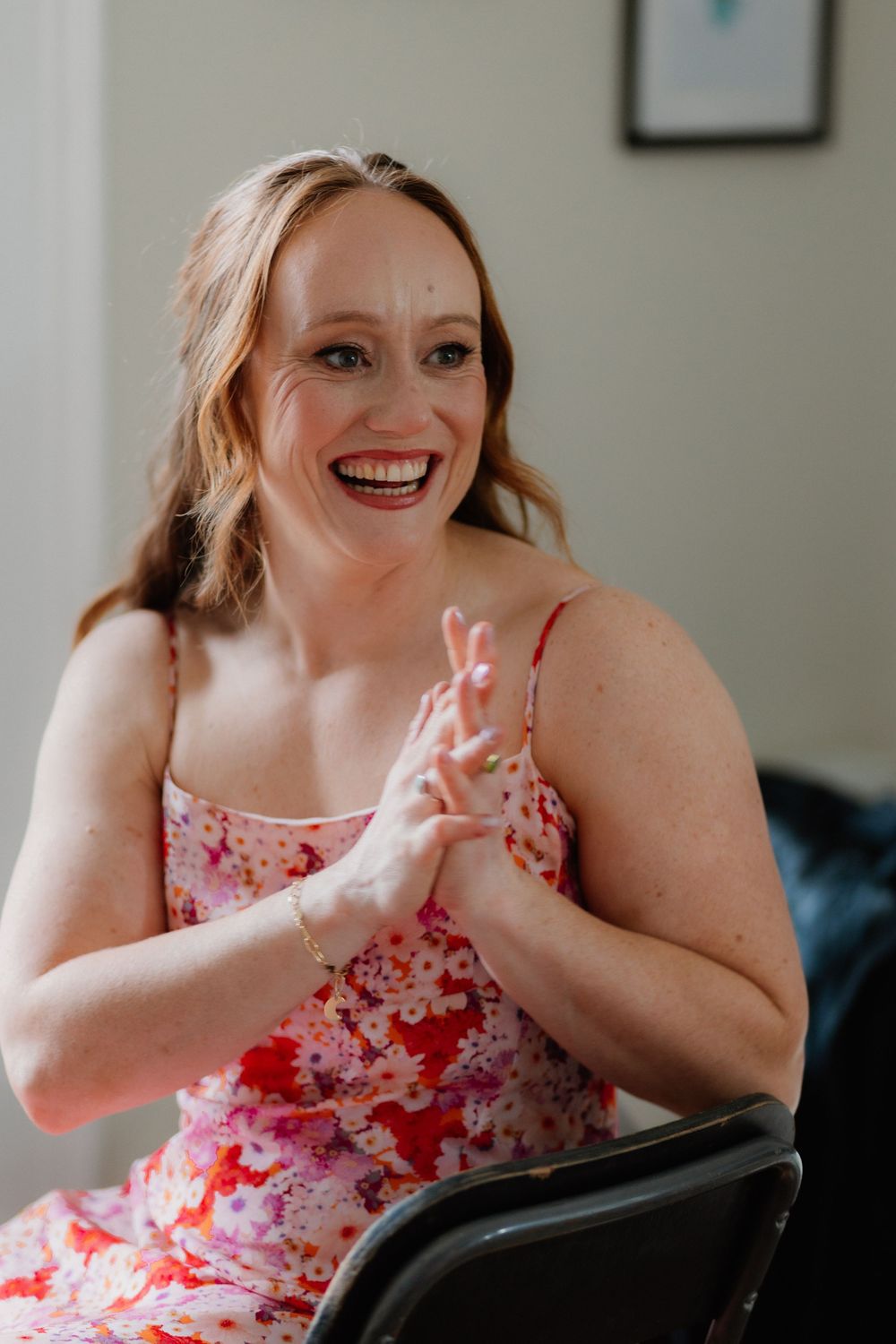 A smiling woman in a floral pink dress sits clapping her hands, laughing joyfully indoors.