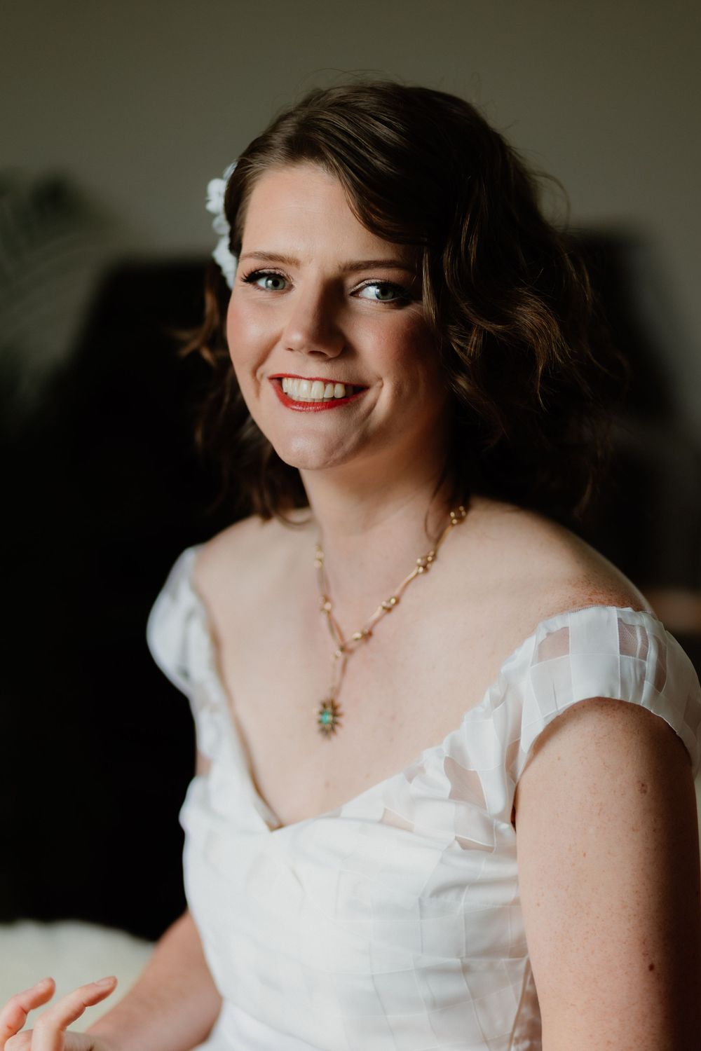 Smiling bride in white off-shoulder gown with gold necklace and floral hair accessory on her wedding day.