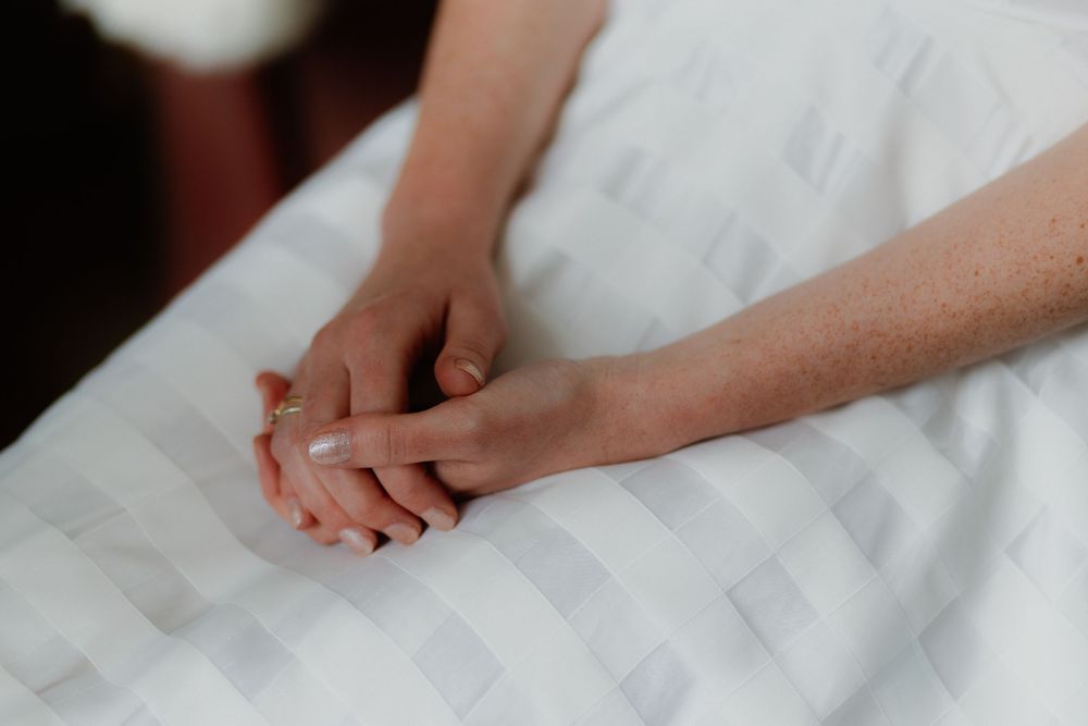 Two people holding hands tenderly on a white bed, wearing a gold ring, symbolizing love and connection.